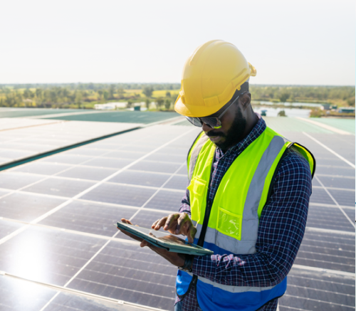 man with a hard hat and vest in the foreground looking at a tablet. A commercial rooftop solar installation in the background.